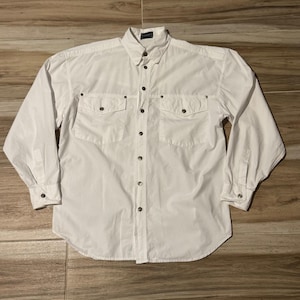 May include: A long-sleeved white button-down shirt featuring a classic collar, two chest pockets with button closures, and a front placket with metallic buttons. The shirt includes a small rectangular label at the neckline and buttoned cuffs.