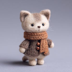 May include: A handcrafted felt cat figurine, featuring a brown and cream color scheme. The cat wears a brown sweater with two buttons and a matching scarf. The cat has a sweet expression with black eyes and white whiskers.