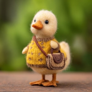 May include: A handcrafted, yellow duckling figurine wearing a yellow sweater with brown polka dots and a small brown satchel. The duckling has an orange beak and is standing on a wooden surface against a blurred green background.
