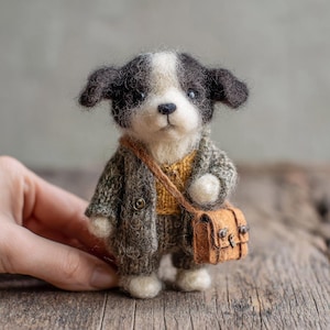 May include: A handcrafted felted dog figurine, featuring a black and white head, wearing a gray coat over a yellow sweater, and carrying a brown satchel. The dog has white paws and is standing on a wooden surface.