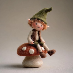 Puede incluir: Una muñeca elfo de fieltro hecha a mano con cabello rojo, mejillas rosadas y un sombrero verde, sentada sobre una seta roja y blanca con lunares. El elfo lleva un chaleco marrón y pantalones verdes, creando un artículo decorativo de temática de cuento de hadas.