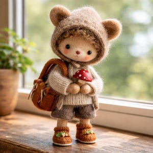 Pode incluir: Boneca de feltro feita à mão usando um capuz de urso marrom, suéter, shorts e botas. A boneca carrega uma mochila marrom e segura um cogumelo vermelho e branco. A boneca está em uma superfície de madeira.