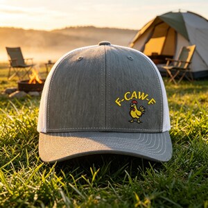 May include: A gray trucker hat with a white mesh back and a curved brim. The hat features the text "F-CAW-F" in yellow above a cartoon chicken. The hat is set against a camping scene with a tent and campfire.