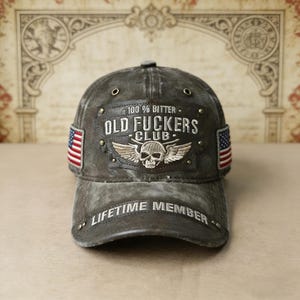 Op de afbeelding: Verweerde grijze baseballcap met de tekst "100% Bitter Old Fuckers Club Lifetime Member". Voorzien van een schedel met vleugels en Amerikaanse vlagpatches aan de zijkanten. Een robuuste pet in vintage-stijl.