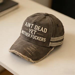 May include: Distressed grey baseball cap with the text "AINT DEAD YET MOTHER FUCKERS" in white. The cap has a curved brim with distressed edges and two grey stripes on the side.