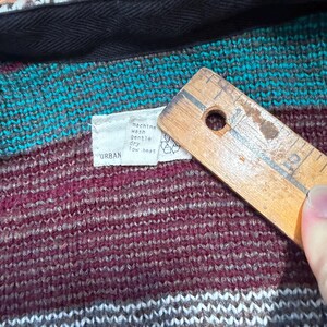 May include: Close-up of a knitted garment with a tag that reads "URBAN" and care instructions. The garment features a color block design with teal, burgundy, and gray stripes. A wooden ruler is partially visible.