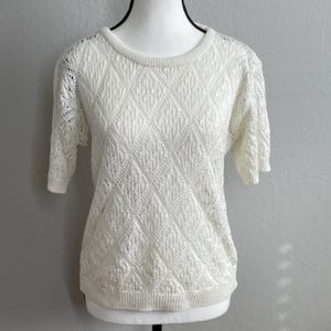 May include: Off-white short-sleeve sweater with a textured diamond pattern. The sweater has a crew neck and ribbed cuffs and hem. The sleeves are a cap sleeve style.