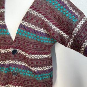 May include: A brown cardigan sweater with a V-neck and two black buttons. The sweater features a geometric pattern in teal, purple, white, and burgundy. The sleeves are long and the knitwear is thick.