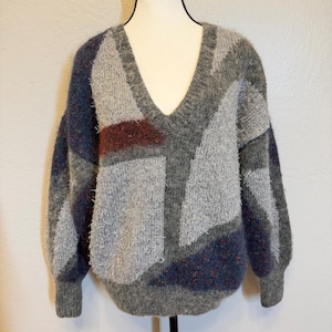 May include: A gray, blue, and burgundy V-neck sweater with a geometric pattern. The sweater has long sleeves and a relaxed fit. The fabric appears to be a soft, textured knit.