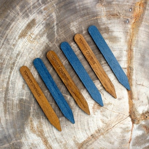 May include: Six wooden plant markers with a pointed end. The markers are in two colours: brown and blue. Each marker has the text "Tommy Owens" engraved on it.