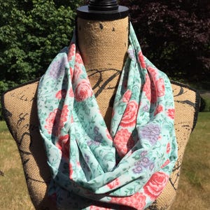 May include: A floral infinity scarf in shades of light blue, pink, and purple. The scarf features a repeating pattern of roses and other flowers. The scarf is displayed on a mannequin.