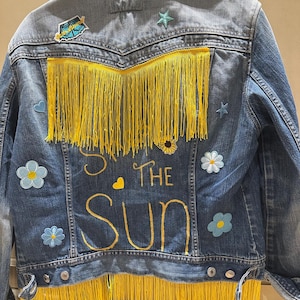May include: A blue denim jacket with yellow fringe and embroidered text reading "So the Sun." The jacket features patches of flowers, stars, and a small bus. Yellow fringe adorns the back and bottom hem of the jacket.