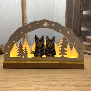 May include: A wooden decorative piece featuring two dog silhouettes, a forest scene, and star cutouts. The arch-shaped design has a warm, yellow glow from the internal lighting. The base is rectangular, and the piece is set on a wooden surface.
