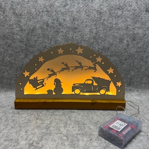 Puede incluir: Decoración navideña de madera con un interior cálido y brillante. El diseño presenta a Papá Noel en su trineo, renos, un muñeco de nieve y un camión que transporta un árbol de Navidad. La decoración está iluminada por pequeñas luces y tiene un paquete de baterías.