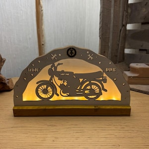 May include: A wooden, illuminated motorcycle silhouette with the word "Samson" and "DDR" on the arch. The motorcycle is a detailed cutout, and the light shines through the design. The base is a dark brown, and the light is a warm yellow.