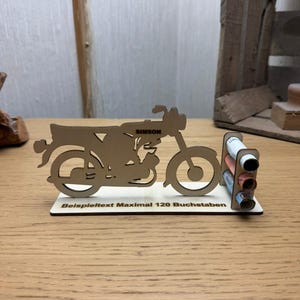 Könnte beinhalten: Ein lasergeschnittenes Motorradmodell aus Holz mit dem Namen "SIMSON". Das Modell steht auf einem Holzsockel mit einem kleinen Halter für gerolltes Papier. Der Sockel trägt den Text "Beispieltext Maximal 120 Buchstaben."