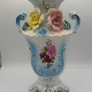 May include: A white ceramic vase with a light blue and gold trim. The vase features three-dimensional yellow, pink, and blue flowers. The vase has a floral design and two curved handles.