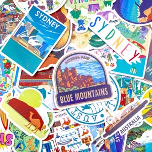 Peut inclure: Une collection d'autocollants en vinyle colorés sur le thème des voyages en Australie, incluant l'Opéra de Sydney, les Blue Mountains et diverses illustrations de faune. Les autocollants affichent des textes comme Sydney, Australia et Blue Mountains.