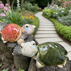 May include: Two ceramic turtle figurines with glossy finishes. One has a red-orange shell, the other a dark green shell. Both have light-colored heads and are perched on a weathered tree stump, set against a garden path.
