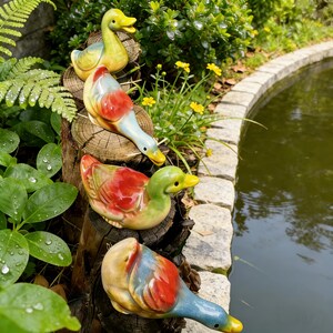 May include: Four ceramic duck figurines in various colors, including yellow, red, green, and blue, are arranged on a tree stump. The ducks are positioned near a pond with a stone border, surrounded by greenery and yellow flowers.
