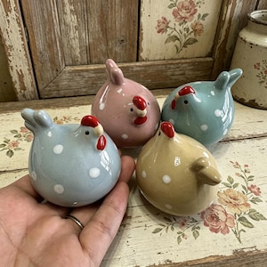 Op de afbeelding: Vier keramische kippenbeeldjes in pastelkleuren: blauw, roze, groen en beige. Elke kip heeft witte stippen, een rode kam en een snavel. De kippen staan op een oppervlak met bloemenpatroon.