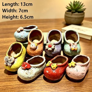 Puede incluir: Una colección de ocho pequeñas macetas de cerámica con forma de zapato en varios colores, incluyendo amarillo, naranja, morado, verde, rojo y rosa. Cada maceta presenta un adorno decorativo como una mariposa, una flor o una cara de animal. Longitud: 13cm, Anchura: 7cm, Altura: 6,5cm.