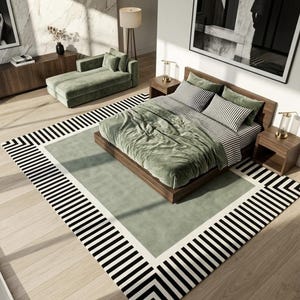 May include: A large rectangular area rug featuring a central sage green field bordered by a wide white band and an outer frame of bold black and white stripes. The rug is positioned under a wooden bed frame with sage green bedding and pillows.