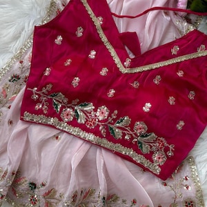 Puede incluir: Una blusa de color magenta con ribete dorado y bordados florales, combinada con una tela transparente rosa claro. La blusa presenta un cuello en V y está adornada con lentejuelas doradas. La tela transparente tiene bordados florales a juego.