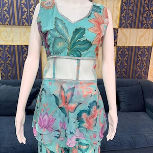Peut inclure: Une tenue turquoise sans manches &agrave; motifs floraux. Le haut a un col en V et une bordure argent&eacute;e. La jupe a des volants superpos&eacute;s. Le motif floral comprend des feuilles et des fleurs roses, violettes et vertes. La tenue est pr&eacute;sent&eacute;e sur un mannequin.