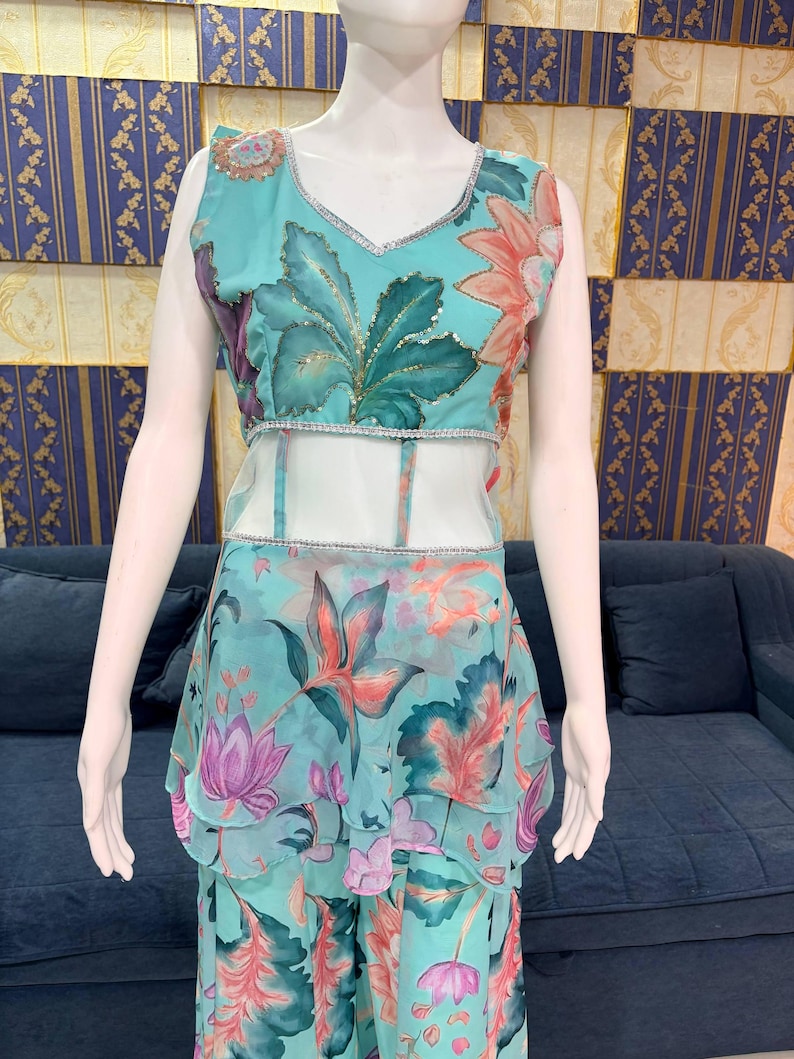 Peut inclure: Une tenue turquoise sans manches &agrave; motifs floraux. Le haut a un col en V et une bordure argent&eacute;e. La jupe a des volants superpos&eacute;s. Le motif floral comprend des feuilles et des fleurs roses, violettes et vertes. La tenue est pr&eacute;sent&eacute;e sur un mannequin.