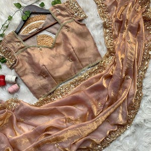 Puede incluir: Un conjunto de sari y blusa color melocotón. La blusa tiene mangas cortas y un escote cuadrado, adornado con lentejuelas doradas. El sari presenta un borde de lentejuelas doradas y está drapeado sobre una superficie blanca.