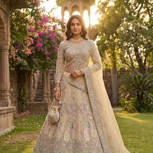 May include: A beige embroidered lehenga with a matching long-sleeved blouse and dupatta. The lehenga features intricate floral and peacock designs with pink and green accents. A small matching purse is held in the hand.