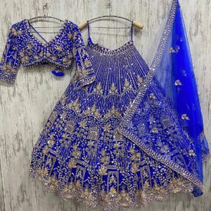 May include: A royal blue lehenga choli set, featuring a sleeveless top, a long skirt, and a matching dupatta. The set is adorned with gold embroidery and sequins. The intricate detailing and rich colour create a luxurious and festive look.
