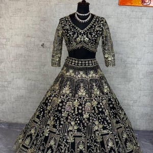 May include: Black and gold embroidered formal gown with a matching crop top and long sleeves. The gown features intricate gold floral and geometric patterns. A beaded necklace is on the mannequin's neck.
