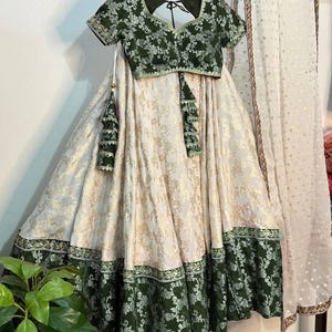 May include: An Indian-style outfit featuring a green floral patterned crop top with short sleeves and a matching skirt. The skirt is cream-colored with gold floral accents and a green floral border. A white scarf with gold dots hangs on the side.