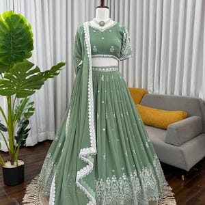 Op de afbeelding: Saliegroene formele jurk met ingewikkelde witte borduursels en een bijpassende dupatta. De jurk heeft een crop top, een zwierige rok en een delicate witte kanten rand. Een ketting siert de mannequin.