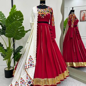 May include: A red and white traditional Indian outfit displayed on a mannequin. The outfit includes a long skirt with gold trim, a cropped top with colorful embroidery, and a matching shawl. The outfit is displayed in front of a mirror.