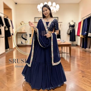 以下が含まれることがあります： ネイビーブルーの伝統的なインドの衣装。精巧な刺繍が施されたクロップトップ、流れるようなスカート、お揃いのスカーフが特徴です。衣装には、金と赤のアクセントが付いた装飾的なボーダーがあります。画像には「SRUSH Handcrafted」というテキストも含まれています。