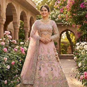 Op de afbeelding: Een lichtroze geborduurde lehenga met een bijpassende blouse met lange mouwen en dupatta. De lehenga heeft pauwen- en bloemborduurwerk. De vrouw draagt gouden sieraden en staat in een tuin met roze bloemen.