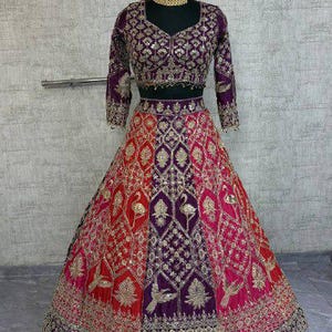 May include: An ornate Indian lehenga in red, pink, and purple, with gold embroidery. The skirt has a detailed pattern with bird designs. The matching top is purple with long sleeves and gold embellishments. A gold necklace is on the mannequin.