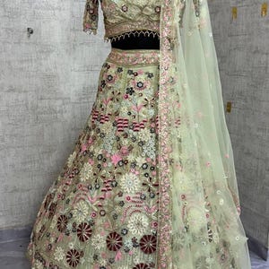 May include: An intricately embroidered, light green lehenga set. The ensemble features a cropped top and a flowing skirt, both adorned with floral patterns in pink, white, and maroon. A matching sheer dupatta and a beaded necklace complete the look.