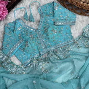 Puede incluir: Una blusa bordada azul claro y tela transparente a juego. La blusa tiene cuello en V y mangas largas, con intrincados bordados florales plateados. La tela transparente está drapeada debajo. El conjunto se exhibe sobre una superficie blanca y esponjosa.