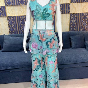 Peut inclure: Une tenue turquoise &agrave; imprim&eacute; floral sur un mannequin. La tenue comprend un haut sans manches avec un col en V et un pantalon large assorti. Le motif floral pr&eacute;sente des accents roses, oranges et verts. Le haut a un panneau transparent &agrave; la taille.