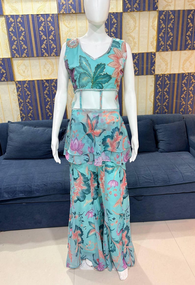 Peut inclure: Une tenue turquoise &agrave; imprim&eacute; floral sur un mannequin. La tenue comprend un haut sans manches avec un col en V et un pantalon large assorti. Le motif floral pr&eacute;sente des accents roses, oranges et verts. Le haut a un panneau transparent &agrave; la taille.