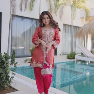 Puede incluir: Una mujer con un salwar kameez color coral con bordados intrincados y pantalones fucsia a juego. Sostiene un pequeño bolso adornado. El atuendo se combina con sandalias. El fondo incluye una piscina y palmeras.