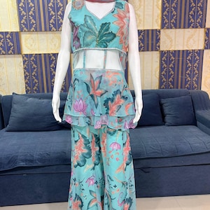 Peut inclure: Une tenue &agrave; imprim&eacute; floral turquoise sur un mannequin. La tenue comprend un haut sans manches avec un col en V, un milieu du corps transparent et un basque &agrave; volants. Un pantalon large assorti et une &eacute;charpe compl&egrave;tent l'ensemble. Le motif floral pr&eacute;sente des accents roses, oranges et verts.