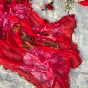 Puede incluir: Una bufanda roja vibrante con grandes diseños florales en tonos rosa y marrón. Los bordes están adornados con cuentas doradas. La bufanda está drapeada sobre una superficie blanca y esponjosa, con algunos carretes de hilo y flores en el fondo.