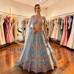 以下が含まれることがあります： 精巧な刺繍と装飾が施された、水色のレヘンガ。お揃いのチョリとドゥパッタが含まれています。画像には「SRUSH Handera」の文字も表示されています。