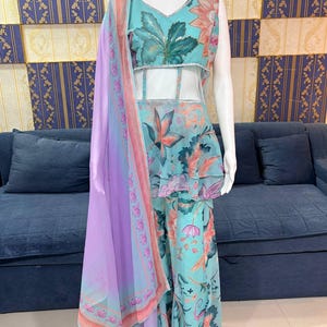 Peut inclure: Une tenue &agrave; imprim&eacute; floral turquoise sur un mannequin. La tenue comprend un haut sans manches avec un col en V, un haut de style p&eacute;plum et un pantalon large. Une &eacute;charpe transparente lavande avec une bordure florale est drap&eacute;e sur la tenue. Le fond pr&eacute;sente un canap&eacute; bleu et un mur &agrave; motifs.