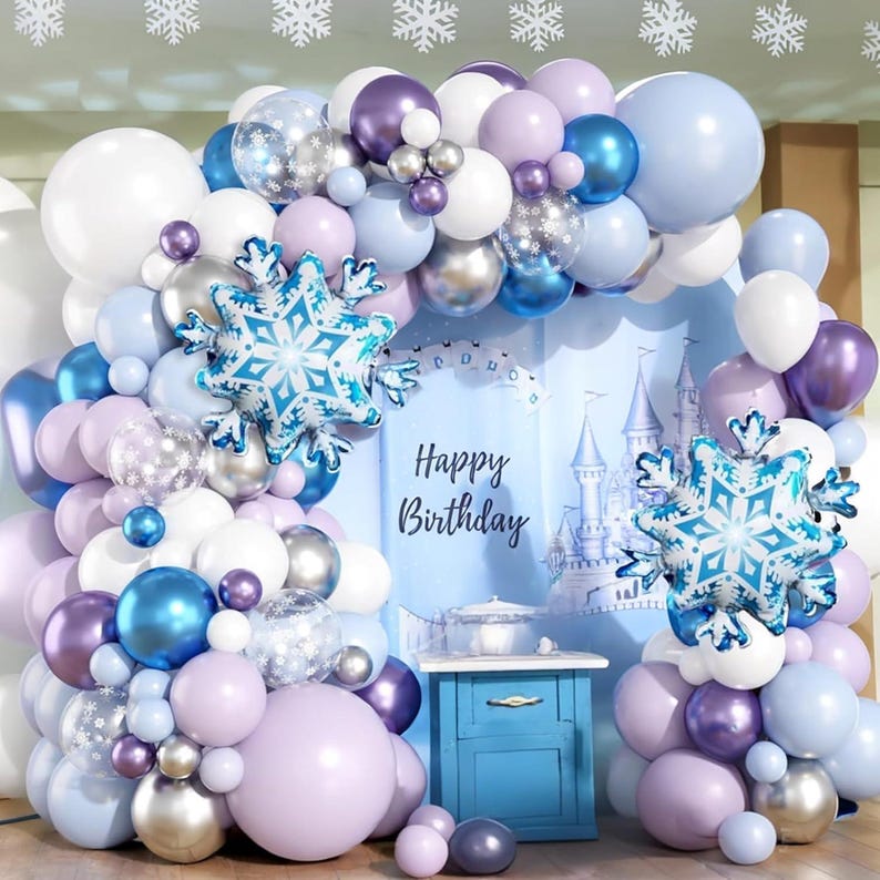 Puede incluir: Un arco de globos festivo para una fiesta de cumplea&ntilde;os, en tonos blancos, azules, plateados y morados. El arco presenta globos de papel de aluminio con forma de copo de nieve y un tel&oacute;n de fondo con un castillo y la frase "Happy Birthday".